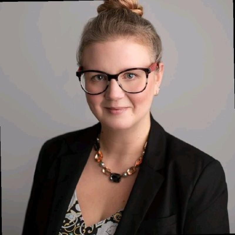 Headshot of white woman with her hair in a bun and wearing a black blazer and red glasses